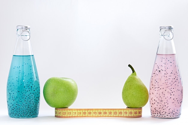 A centimeter, an apple, a pear and glass bottles with pink and blue basil seed stand on a white background