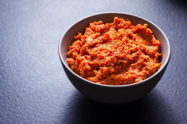 Ceramic bowl with cashew dipping sauce of bright orange colour Thick dip with red paprika ready to be served with vegetables or grilled meat Spicy snack closeup on dark grey background