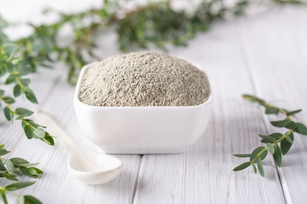 Ceramic bowl with green clay powder and fresh eucalyptus leaves on white background. Concept of face and body care.