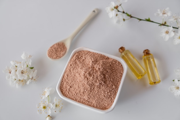 Ceramic bowl with red clay powder, ingredients for homemade facial and body mask or scrub and fresh sprig of flowering cherry on white surface. Spa and bodycare concept.