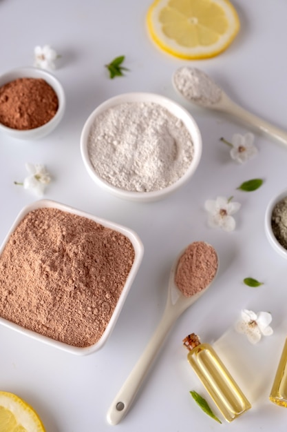 Ceramic bowl with red clay powder, ingredients for homemade facial and body mask or scrub and fresh sprig of flowering cherry on white surface. Spa and bodycare concept.