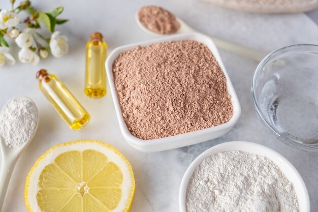 Photo ceramic bowl with red clay powder, ingredients for homemade facial and body mask or scrub and fresh sprig of flowering cherry on white surface. spa and bodycare concept.