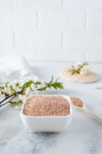 Ceramic bowl with red clay powder, ingredients for homemade facial and body mask or scrub and fresh sprig of flowering cherry on white surface. Spa and bodycare concept.