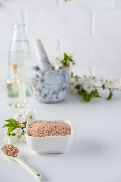 Photo ceramic bowl with red clay powder, ingredients for homemade facial and body mask or scrub and fresh sprig of flowering cherry on white surface. spa and bodycare concept.