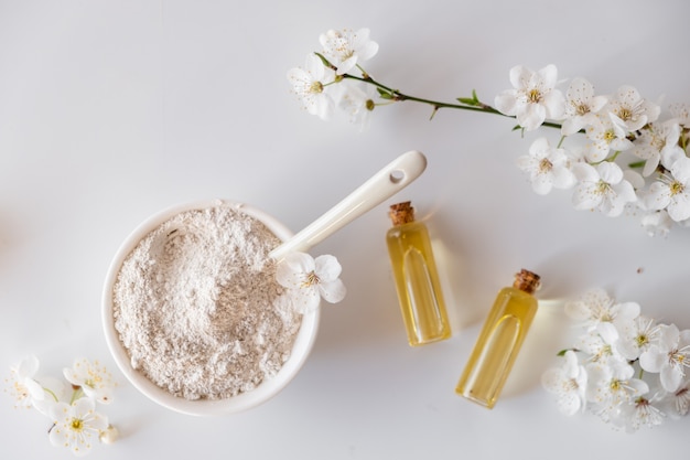 Ceramic bowl with white clay powder on white surface. Ingredients for homemade facial and body mask or scrub and fresh sprig of flowering cherry. Spa and bodycare concept.