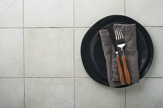 Ceramic empty black plate silverware and linen kitchen towel\
napkin on old ceramic tile table background cooking stone backdrop\
top view with copy space flat lay