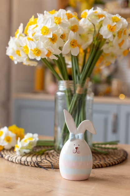 A ceramic figurine of a cute striped hare stands on the kitchen table next to a glass vase with daffodil flowers