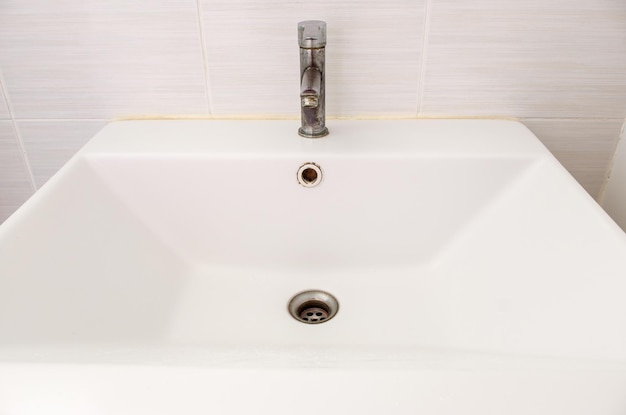 The ceramic sink basin with the stained faucet near the white tile wall of the restroom in the city house front view for the background