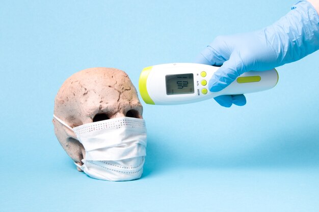 Ceramic skull in mask and a hand in a medical glove with non-contact thermometer blue background, copy space