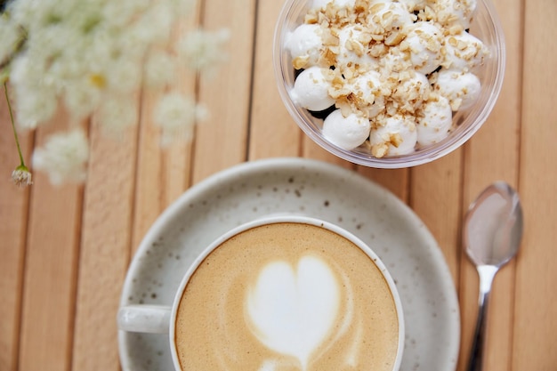 Ceramic white cup of cappuccino with heart vegan portion of nuts dessert in a disposable cup light breakfast or coffee break with blurred foreground of chamomile flowers