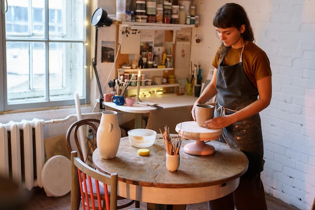 仕事中のスタジオの若い女性の陶器スタジオの所有者のマスタークラスでエプロン成形花瓶の陶芸家