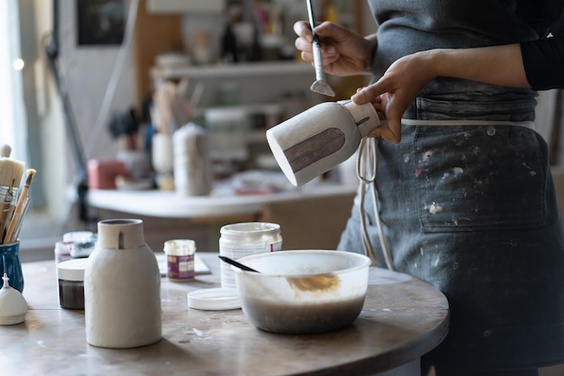 Ceramist in black apron paints clay vase with thick paintbrush near table in cozy workshop