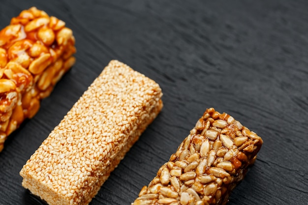 Cereal granola bar with peanuts, sesame and sunflower seeds on a cutting board on a dark stone table. View from above. Three Assorted Bars