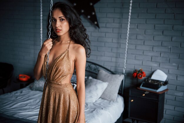 Цепная кровать. Девушка в золотом платье стоит перед белой кроватью в роскошных апартаментах