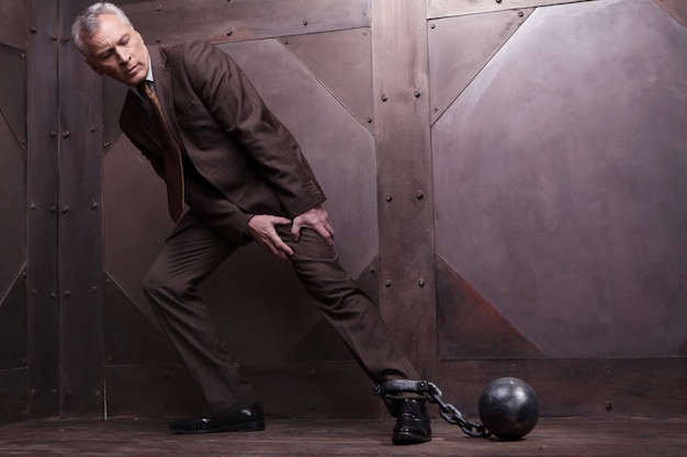 Chains on his leg. Full length of grey hair senior man in formalwear trying to go with the shackles on his leg