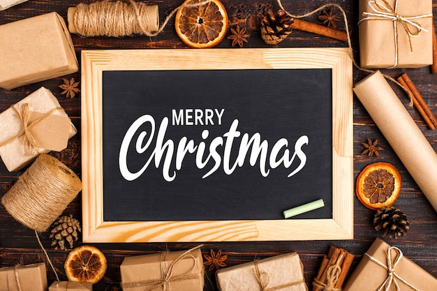 Chalk board in the middle of gifts of dried orange, cinnamon, pine cones, anise on a white table. Merry christmas inscription.