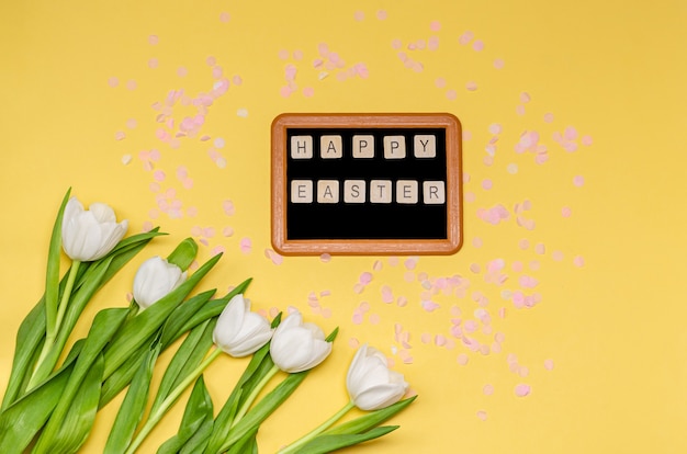 Chalk board with the words Happy Easter, pink decorative paper confetti and a bouquet of white tulips on a yellow wall copy space, flat lay