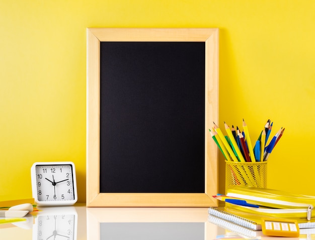 Chalkboard and school supplies on white table by the yellow wall. Side view, empty 