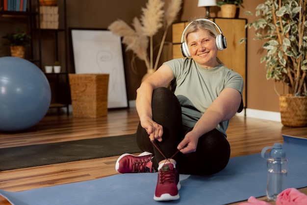 Chaming elderly woman sitting on yoga mat smiling at camera tiing shoelaces having fun