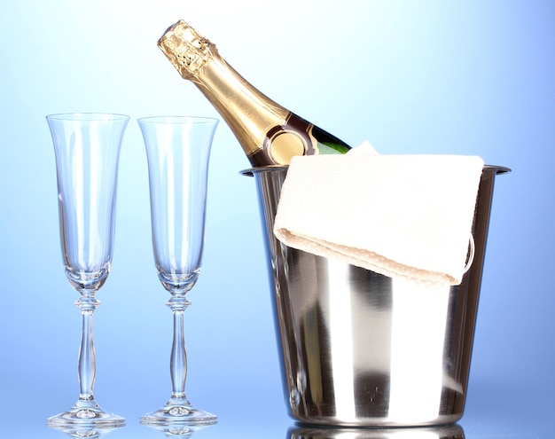 Champagne bottle in bucket with ice and glasses on blue surface