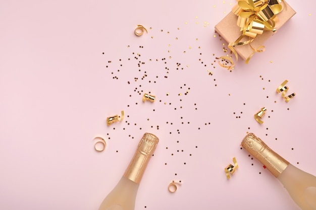 Champagne bottle, gift or present box and golden shiny sparkle star confetti on pink background. Christmas or New Year composition or card. Celebration flat lay. Party creative concept. Top view.