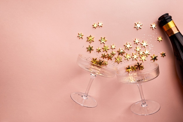 Champagne bottle with golden glittering splashes and champagne glasses on pink background flat lay