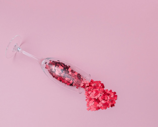 Champagne glass filled with heart shaped confetti and heart made of confetti on the pink background