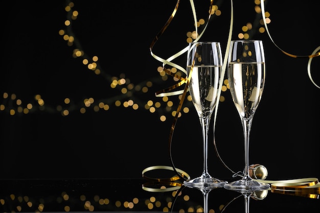 champagne in glasses and party streamers on mirror surface against black background with blurred lights space for text