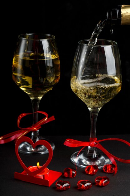 Champagne is poured into glasses on a black surface next to a heart-shaped candlestick with a burning candle and a box with a ring