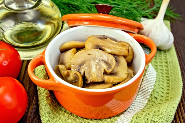 Champignons fried in a red ceramic pot on a napkin, garlic, dill and tomatoes on background dark wooden boards