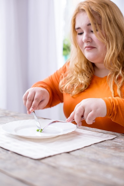 ライフスタイルの変化。テーブルに座ってエンドウ豆を食べる悲しい太りすぎの女性