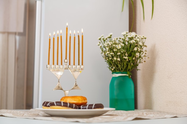 Chanukiah with all burning candles and donuts and a vase of flowers on the table.