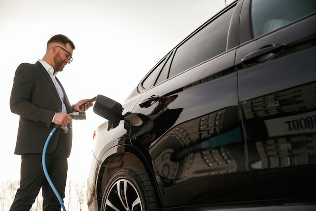 Charging the electric vehicle Businessman in suit is near his black car outdoors