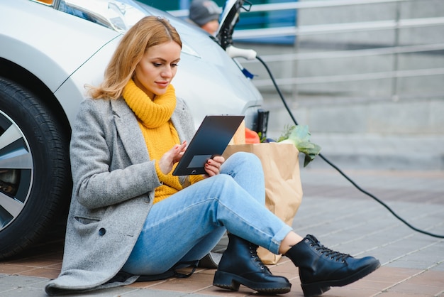 電気ガソリンスタンドで電気自動車を充電します。車のそばの女性。