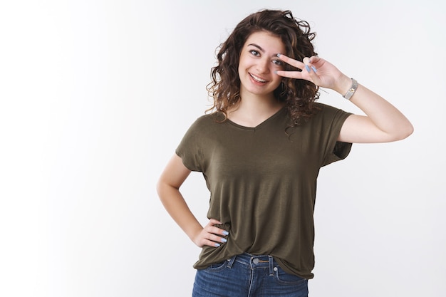 Charismatic lucky cute caucasian woman show victory peace gesture make disco sign near eye having fun good day standing positive happy smiling white teeth near studio background