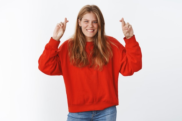 Carismatica e ottimista giovane donna europea in maglione rosso oversize alzando le mani con le dita incrociate per buona fortuna sorridendo ampiamente sentendo fortuna e fortuna dalla sua parte, fede nella vittoria e nel successo