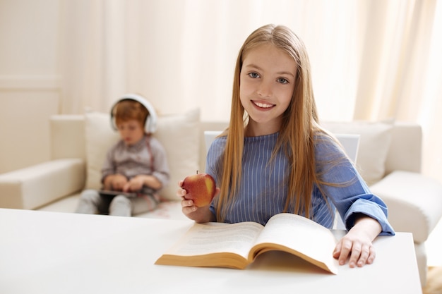 Charismatisch slim schattig kind dat aan tafel zit en iets leest terwijl hij een appel eet als tussendoortje