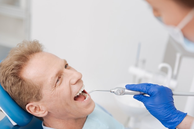 Charismatische knappe man van middelbare leeftijd arts een bezoek brengen en zittend in de stoel van de tandarts tijdens het ondergaan van een professionele procedure