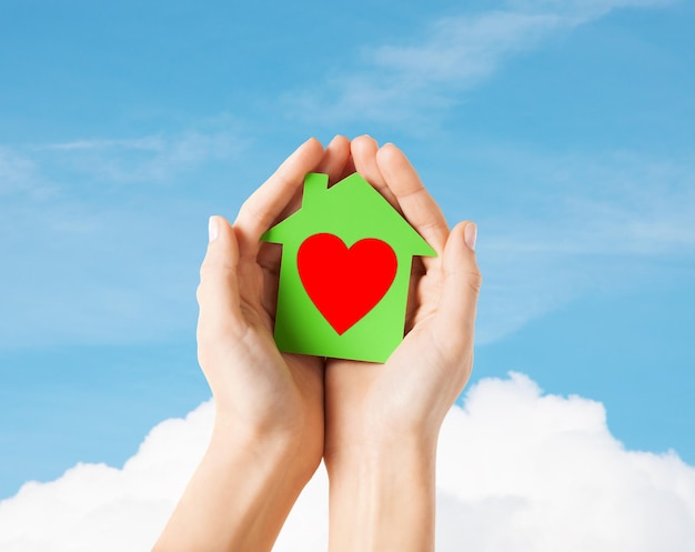 Concetto di casa di beneficenza, immobiliare e famiglia - foto del primo piano delle mani femminili che tengono la casa di carta verde con cuore rosso