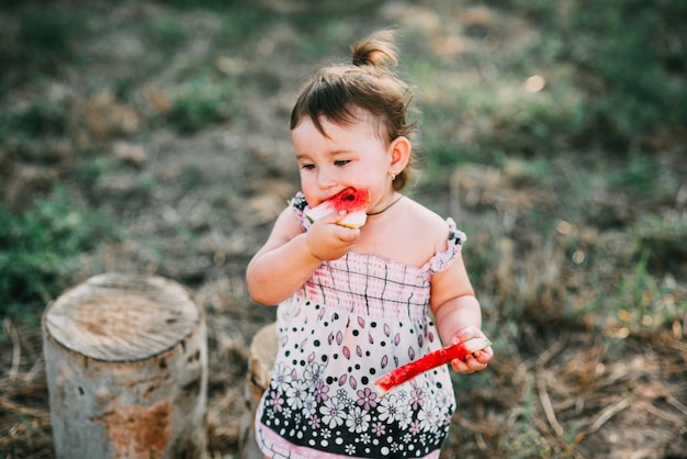 Charmant anderhalf jaar oud meisje dat buiten watermeloen eet, in het dorp tegen de bomen met een staart op zijn hoofd