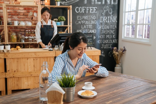 Charmant Aziatisch meisje in streepoverhemd glimlacht en chats in mobiele telefoon. vrouw werknemer aan café tafel met croissant en warme kop koffie met behulp van sms-bericht van de mobiele telefoon. vrouwelijke barista aan het werk in balie