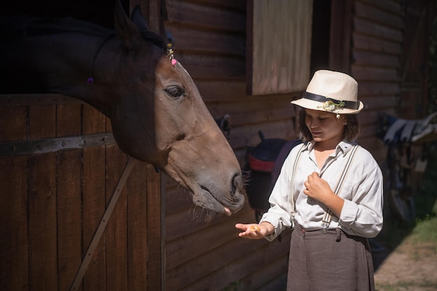 Charmant jong meisje in vintage kleding staande met haar bruine paard dat uit het hoofd van de schuur steekt en...