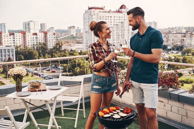 Charmant jong stel in casual kleding die barbecue voorbereidt en glimlacht terwijl ze buiten op het dakterras staan