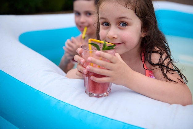 Charmant Kaukasisch meisje drinkt een verse cocktail en glimlacht terwijl ze naar de camera kijkt terwijl ze plezier heeft in een opblaasbaar zwembad