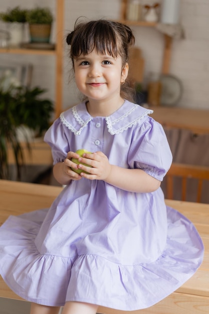 charmant klein meisje in een lila jurk eet een groene appel in de keuken. ruimte voor tekst, banner