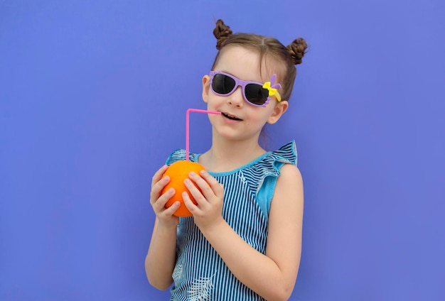 Charmant meisje in zomerglazen drinkt sinaasappelsap op een paarse achtergrond Gezonde levensstijl Kopieer ruimtes