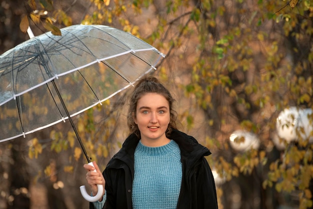 Charmant meisje met blauwe ogen kijkt in de camera in het herfstpark met transparante paraplu Walk in park