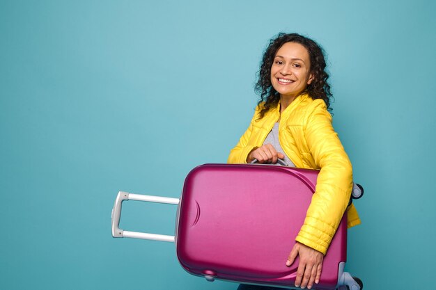 Charmante Afro-Amerikaanse vrouw reiziger in fel geel jasje, glimlachend met vrolijke brede glimlach camera kijken en haar koffer onder de armen houden, geïsoleerd op blauwe achtergrond met kopie ruimte