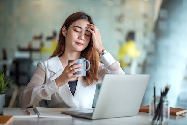 Charmante Aziatische vrouwelijke beambte die op een laptop computer werkt en geniet van het drinken van koffie in een modern kantoor