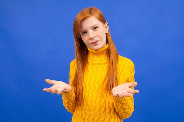 Charmante blanke roodharige meisje in een gele trui zwaaien met haar armen en twijfels op blauw